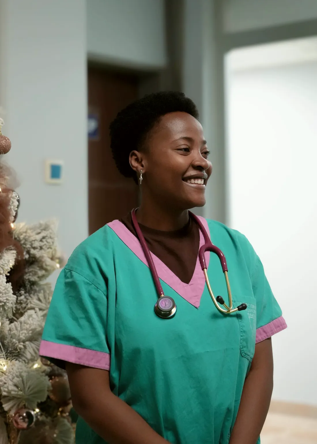 A smiling physician assistant in scrubs with a pink stethoscope, standing in a hospital hallway.